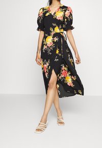 Kvinna klädd i svart midi omslagsklänning med gul och rosa blommig mönstring och puffärmar, i kombination med vita strappy klackskor.