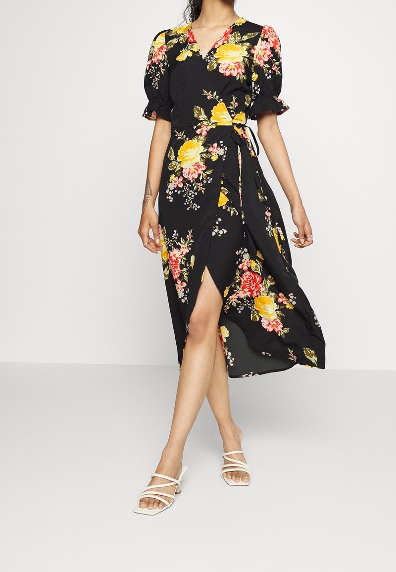Kvinna klädd i svart midi omslagsklänning med gul och rosa blommig mönstring och puffärmar, i kombination med vita strappy klackskor.