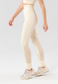 Leggings color beige chiaro con texture a coste, pannelli in rete e ritagli, abbinati a scarpe da corsa viola chiaro.