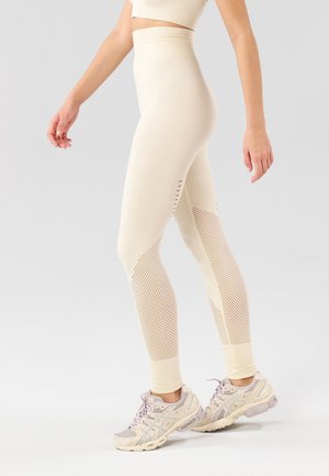 Leggings color beige chiaro con texture a coste, pannelli in rete e ritagli, abbinati a scarpe da corsa viola chiaro.