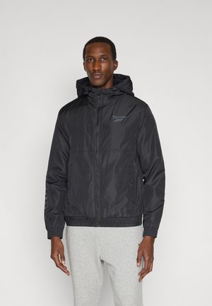 Mann trägt schwarze Reebok-Kapuzenjacke mit Reißverschluss und hellgraue Jogginghose, steht vor schlichtem hellem Hintergrund.