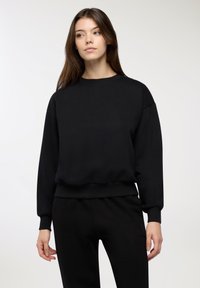 Giovane donna con lunghi capelli castani che indossa una semplice felpa nera a maniche lunghe e pantaloni neri su uno sfondo bianco.