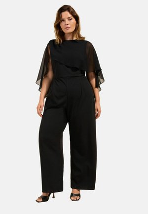 Kvinde iført en sort jumpsuit med gennemsigtige kappeærmer og bukser med vide ben, parret med sorte åbne hæle, stående mod en ensfarvet baggrund.