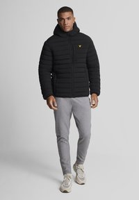 Zwarte gewatteerde jas met capuchon, voorzien van een rits aan de voorkant en een geel logo. Gecombineerd met grijze joggingbroek en beige sneakers. Soepele textuur.