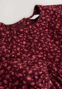 Bordeauxfarbenes Oberteil mit Rundhalsausschnitt, das ein pink- und gelbfarbenes Leopardenmuster und ein strukturiertes Gewebe aufweist. Mit Rüschensaum akzentuiert.