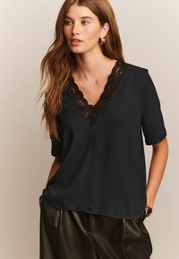 Haut noir à manches courtes avec un décolleté en V, orné d'une bordure en dentelle à volants. Tissu doux, coupe décontractée et coutures discrètes. Porté avec un pantalon sombre.