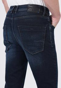 Homme portant un jean en denim bleu foncé avec des poches arrière et une étiquette de marque en cuir noir sur la ceinture, la main dans la poche avant.