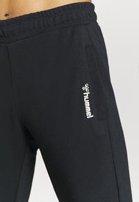 Schwarze Sporthose mit elastischem Bund, ausgestattet mit einer Seitentasche und einem weißen "hummel"-Logo am unteren Bein. Glatte Stoffstruktur.