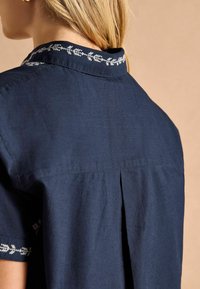 Cămașă bleumarin cu guler brodat și model floral, mâneci scurte și pliuri în spate pentru un plus de mobilitate și confort.