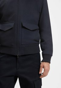 Navy zip-up jacket with two flap front pockets, ribbed hem and cuffs, made from a soft, textured fabric, paired with dark pants.