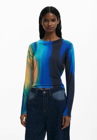 Long-sleeve knit top featuring blue, green, and orange abstract pattern, with a round neck and ribbed hem, paired with dark, textured jeans.