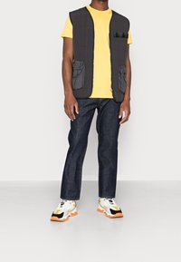 Chaleco acolchado negro con dos bolsillos frontales, camiseta amarilla debajo, jeans de denim oscuro y zapatillas blancas con detalles en negro y naranja.