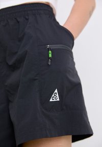 Black shorts featuring a textured fabric, elastic waistband, side pocket with green zipper pull, and a white triangular logo on the front.