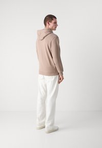 Felpa color crema con cappuccio e polsini a costine, abbinata a pantaloni bianchi e sneakers chiare, vista da dietro su uno sfondo neutro.