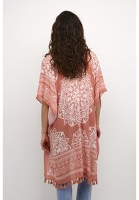 Lyserød-orange kaftan med intrikate hvide mandala-mønstre, letvægtsstof, løs pasform og frynset kant, båret over blå jeans.