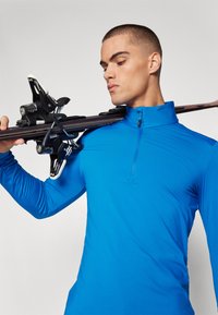 Langermet, blå treningsgenser med halv-zip krage. Stoffet er glatt og tettsittende, med et sømløst design. Skistaver hviler på skulderen.