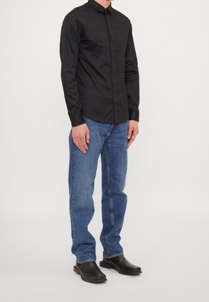 Mann trägt ein schwarzes langärmeliges Hemd mit Knöpfen, blaue Jeans und schwarze Schuhe mit quadratischer Spitze, steht vor weißem einfachem Hintergrund.