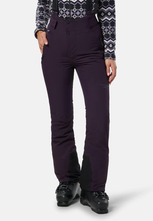 Femme portant des chaussures de ski noires, un pantalon de ski violet avec des bretelles et un haut à manches longues à motifs, debout avec une main dans la poche.