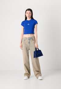 Une jeune femme se tient devant un fond clair, portant un top bleu court et un pantalon beige loose, tenant un petit sac à main bleu.