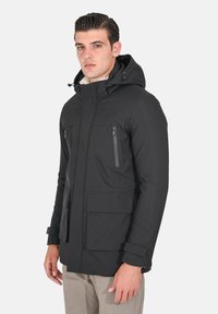 Giacca impermeabile nera con cappuccio, dotata di due tasche frontali con zip, una grande tasca frontale con patta e polsini regolabili. Tessuto liscio.