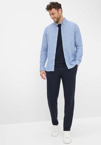 Glunderende man met krullend haar draagt een lichtblauwe overhemd over een zwart T-shirt, marineblauwe broek en witte sneakers, staande met één hand in zijn zak.