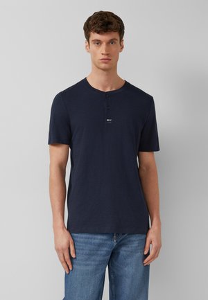 Junger Mann mit navy-blauem Henley-Shirt mit kurzen Ärmeln und blauen Jeans, stehend vor einem schlichten grauen Hintergrund.