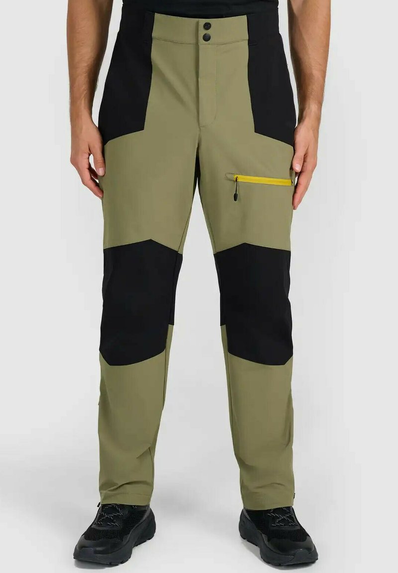 Homme portant un pantalon de randonnée vert olive et noir avec une poche zippée jaune sur la cuisse, debout les mains détendues le long du corps.