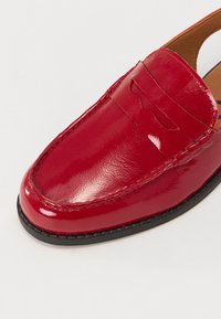 Mocassino in pelle rossa con finitura lucida, caratterizzato da una cucitura superiore e un piccolo dettaglio a taglio. Suola in gomma nera con una leggera texture.