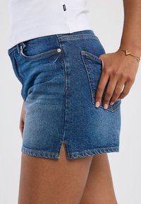 Denim shorts in light blue with yellow stitching, featuring a front pocket, a back patch pocket, and a slit at the hem.