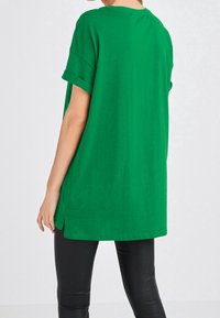 T-shirt verde oversize con maniche arrotolate, tessuto morbido, scollatura tonda e spacchi laterali, abbinato a leggings neri aderenti.