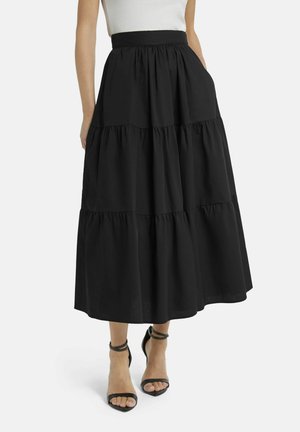 Vrouw draagt een zwarte midi-rok met hoge taille en laagjes, met zakken, gecombineerd met zwarte hakken met enkelbandje en een witte mouwloze top.