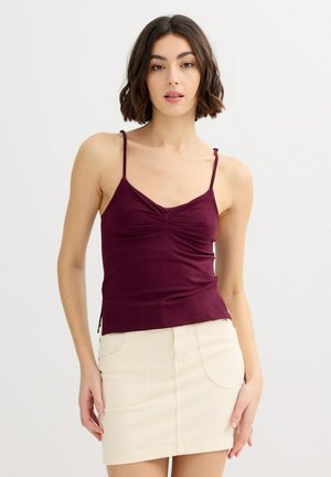 Mujer con cabello corto oscuro, vestida con una blusa sin mangas color burdeos y una minifalda de cintura alta color crema, de pie frente a un fondo claro y liso.