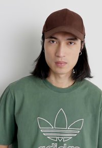 Brączowy czapka baseballowa z gładkiego materiału i okrągłą koroną, w połączeniu z zielonym T-shirtem z okrągłym dekoltem z białym logo.