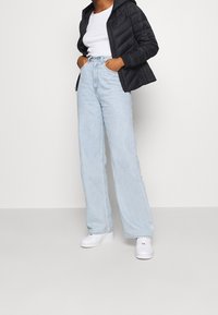 Doudoune noire, t-shirt blanc, jeans taille haute bleu clair, baskets blanches. La doudoune a une fermeture éclair, les jeans sont larges avec une texture délavée.