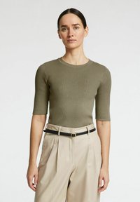 Olivno rebrasto pleteno zgornje oblačilo s kratkimi rokavi, ki ima okrogel izrez in subtilno navpično teksturo. V kombinaciji z visokim pasom bež hlačami in črnim pasom.