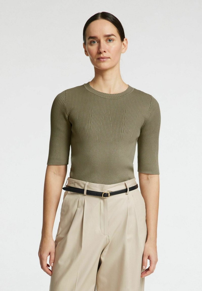 Olivno rebrasto pleteno zgornje oblačilo s kratkimi rokavi, ki ima okrogel izrez in subtilno navpično teksturo. V kombinaciji z visokim pasom bež hlačami in črnim pasom.