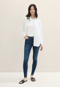 Balta ilgomis rankovėmis palaidinė dėvima ant baltos marškinėlių, mėlyni siauri džinsai ir juodos basutės. Lygi audinio tekstūra ir aptakus dizainas.