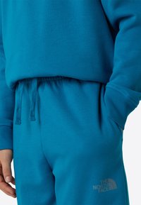 Tyrkysová mikina a tepláky z mäkkej bavlnenej zmesi. Majú rebrované manžety, sťahovací pás a logo na nohavici. Hladká textúra.