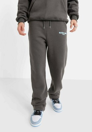 Graue Sweatpants mit einer lockeren Passform, ausgestattet mit einem verstellbaren Kordelzug und einem kleinen blauen Logo. Kombiniert mit blau-weißen Sneakers.