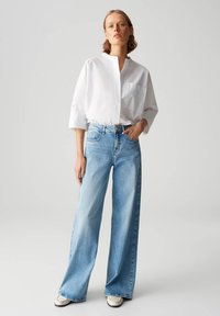 Chemise blanche à col avec une poche poitrine, associée à un jean bleu clair à taille large. Le modèle porte des chaussures blanches. Look simple et décontracté.