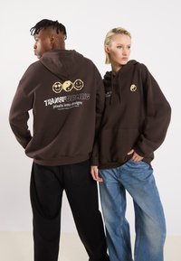 Brown hoodies with graphic designs including smiley faces and yin-yang symbols. The material appears soft, with a relaxed fit and pouch pockets.