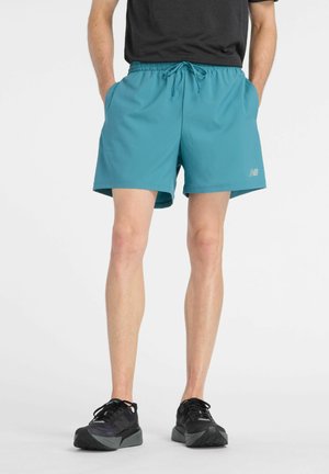 Homme portant un short de sport bleu sarcelle et des chaussures de course noires, debout les mains dans les poches sur un fond blanc.