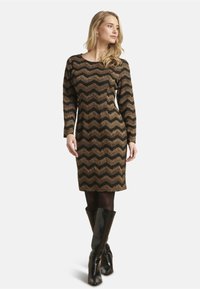 Knielang jurkje in zwart en goud met een zigzagpatroon. Lange mouwen, gemaakt van textuurstof. Gepaard met glanzende zwarte laarzen tot de knie.