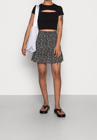 Femme portant un crop top noir, une mini-jupe à fleurs, des sandales à plateforme noires, portant un sac à épaule blanc, se tenant contre un fond uni.