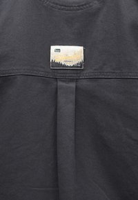 Σκούρο γκρι ύφασμα με ραμμένη ορθογώνια ετικέτα Columbia Sportswear που απεικονίζει δέντρα, βουνά και το κείμενο "Tested Tough" και "Since 1938".