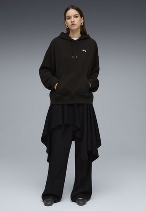 Kobieta w za dużej czarnej bluzie Puma, asymetrycznej czarnej spódnicy nałożonej na szerokie czarne spodnie oraz czarnych butach na szarym tle.