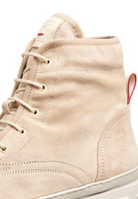 Zapatilla alta de ante beige con cordones beige, ojales dorados y un bucle rojo contrastante en el talón. Suela de goma transparente, textura suave.