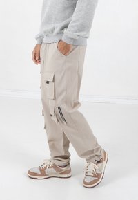 Beige cargobroek met meerdere zakken, ritsen en een relaxte pasvorm, gecombineerd met bruine en grijze sneakers. Grijze sweatshirt zichtbaar bij de taille.