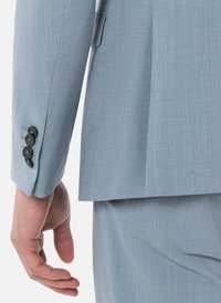 Vaaleansininen puvuntakki, jossa on teksturoitu kangas, kapea istuvuus ja neljä mustaa nappia hihansuissa sekä hillitty laskos takana.