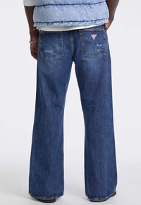 Jeans in denim blu a gamba larga con una finitura sbiadita, tasche posteriori con un logo triangolare rosso e dettagli di cucitura visibili.
